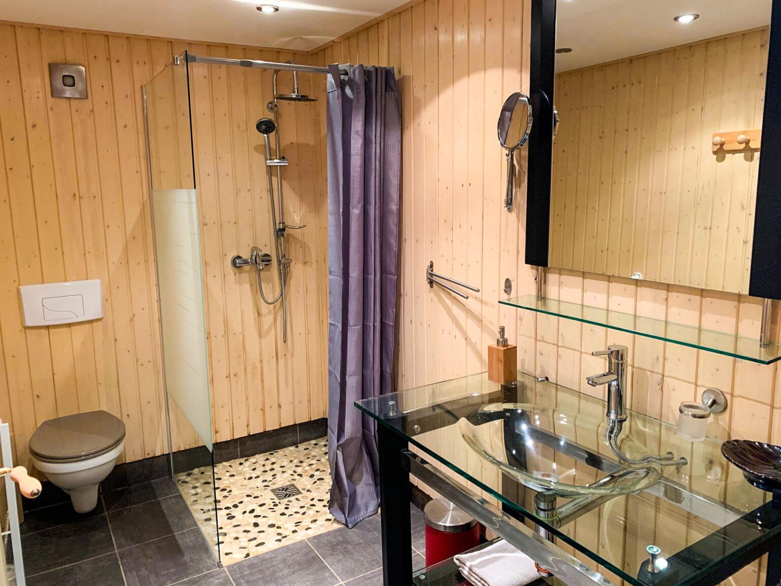 Au coeur des rêves au mont Chateleu salle de bain du gîte. Au premier plan, un lavabo tout en verre transparent, surmonté d'un miroir. En arrère plan une douche à l'italienne avec un sol en galets et à coté de celle ci, des WC.