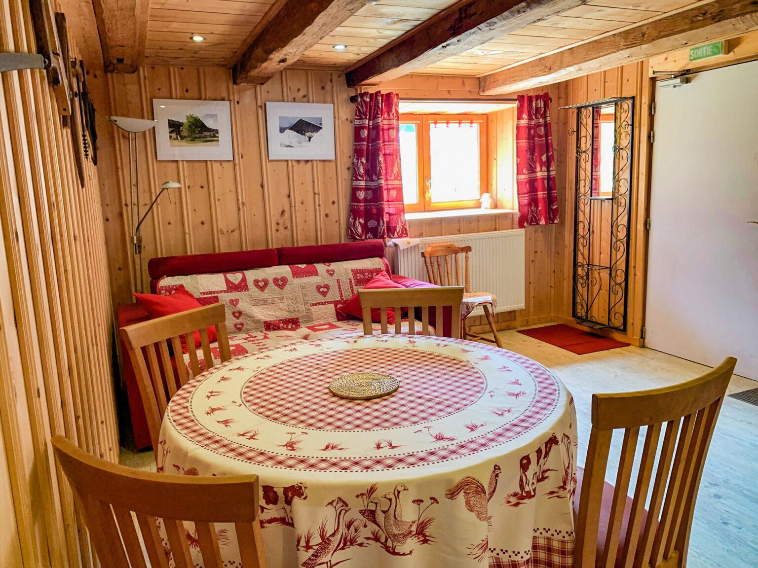 Au coeur des rêves au mont Chateleu détail du gîte, pièce principale contenant une table ronde en premier plan recouverte d'une nappe, avec ses quatre chaises en bois. En arrière plan se trouve un canapé rouge et une chaise en bois est placée à côté de celui-ci.La pièce est tout en bois, ambiance chalet de montagne.