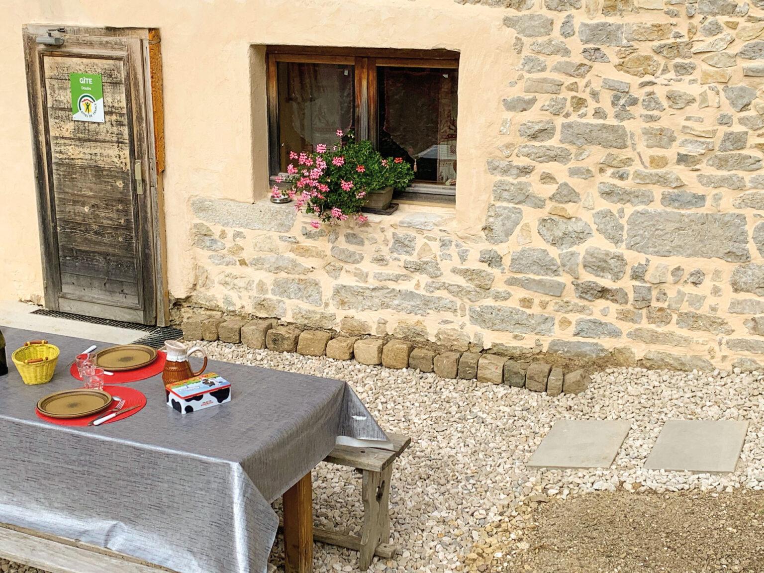 Au coeur des rêves au mont Chateleu détail du gite montrant une table en bois recouverte d'une nappe argentée avec de la vaisselle disposée dessus, la porte et la fenêtre du gite en arrière plan